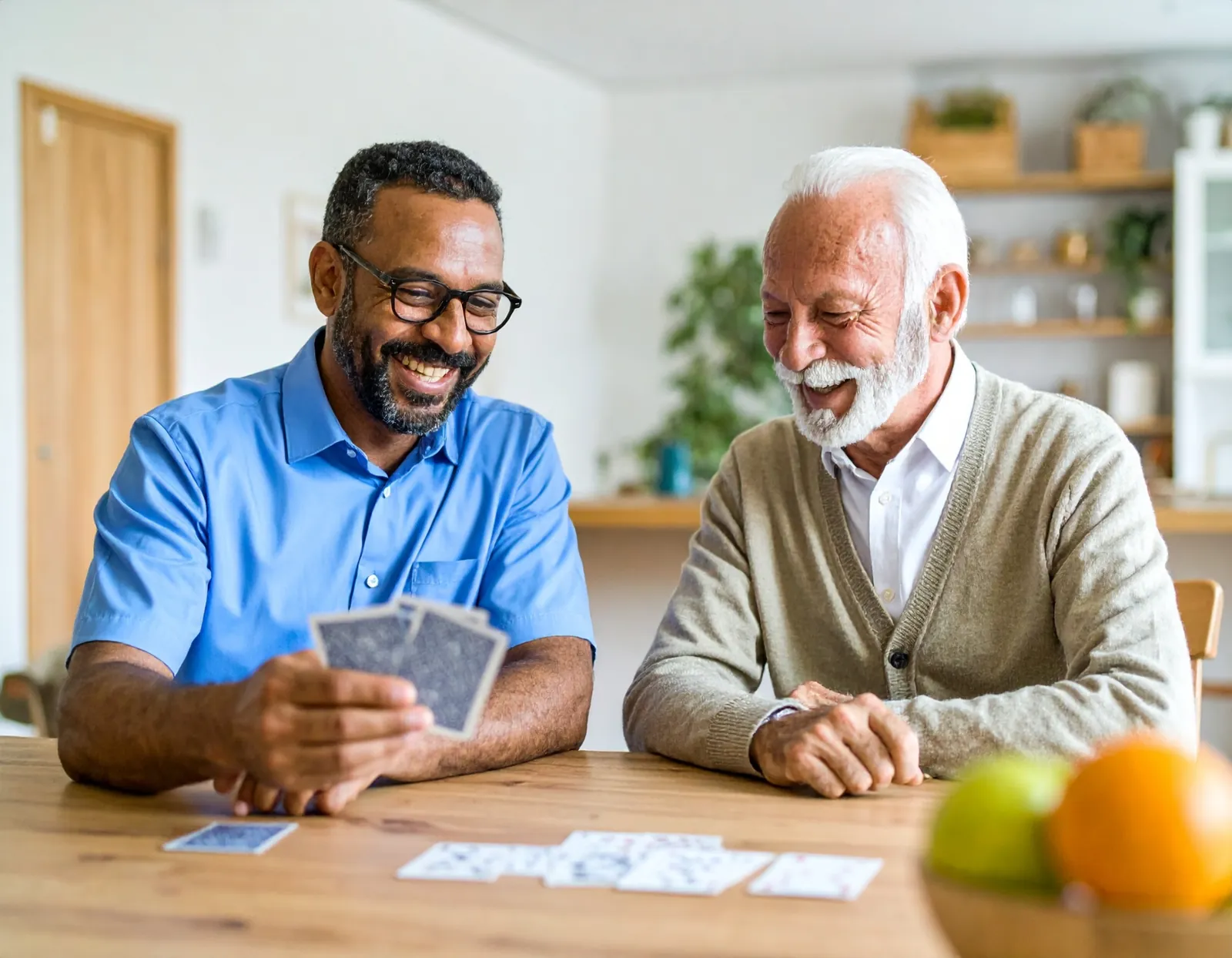 A caregiver and an older man laughing over a game of cards at the kitchen table