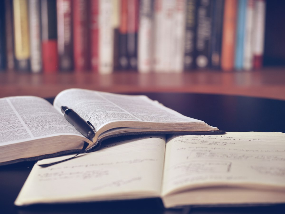 An open notebook and textbooks on a desk in front of a bookshelf